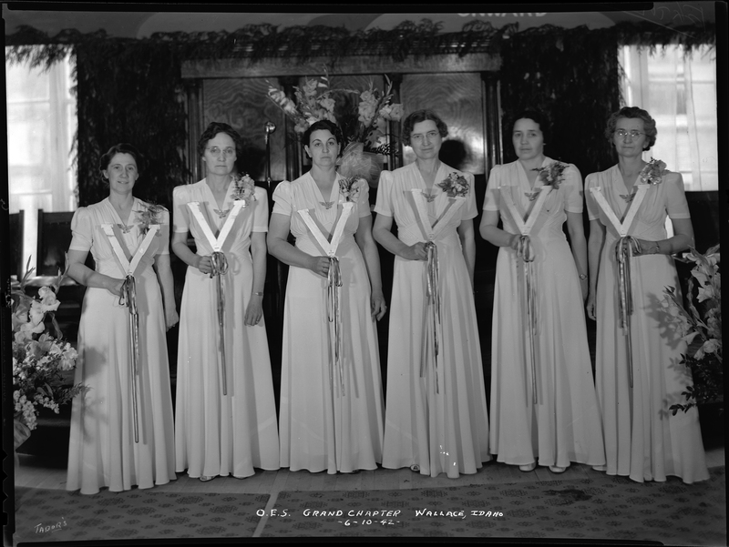 Six women from the Order of Eastern Star Idaho Grand Chapter stand together in matching dresses. They are all holding up something and looking at the camera.