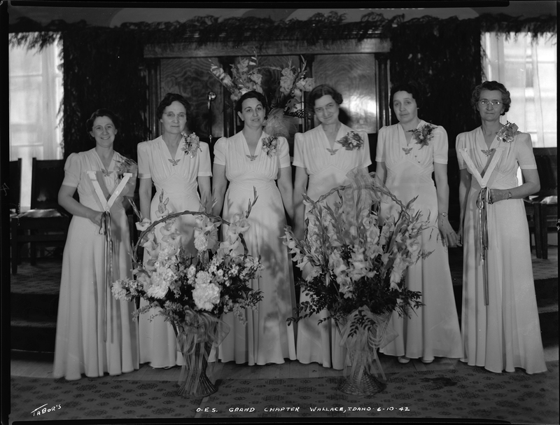 Six women from the Order of Eastern Star Idaho Grand Chapter stand together in matching dresses. They are all looking at the camera with two large bouquets of flowers in front of them.