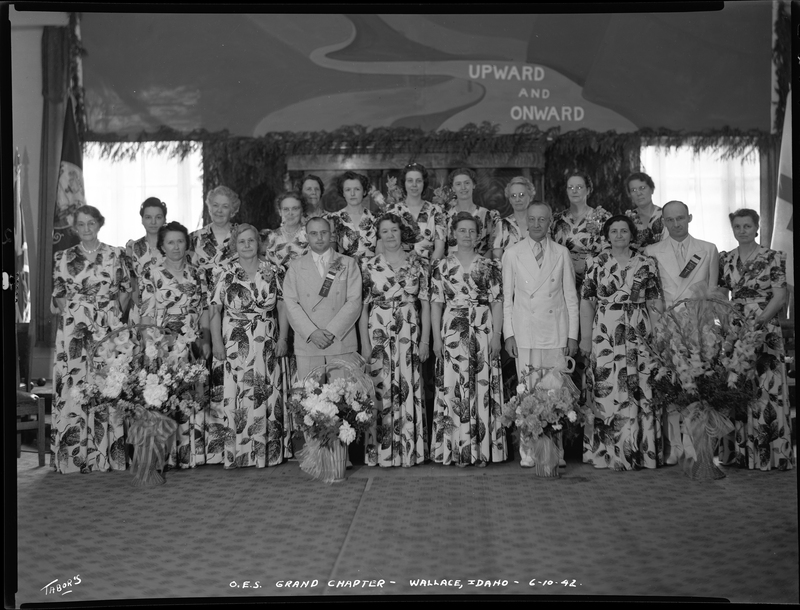 Group of women and men for the Order of Eastern Star Idaho Grand Chapter standing together. The women are all wearing matching dresses and the men are in suits, and there are bouquets of flowers on the ground in front of everyone. A flag can be seen in the corner, and the wall behind everyone reads "upward and onward."