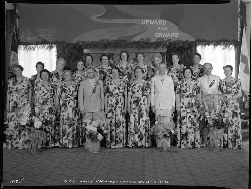 Group of women and men for the Order of Eastern Star Idaho Grand Chapter standing together. The women are all wearing matching dresses and the men are in suits, and there are bouquets of flowers on the ground in front of everyone. A flag can be seen in the corner, and the wall behind everyone reads "upward and onward."