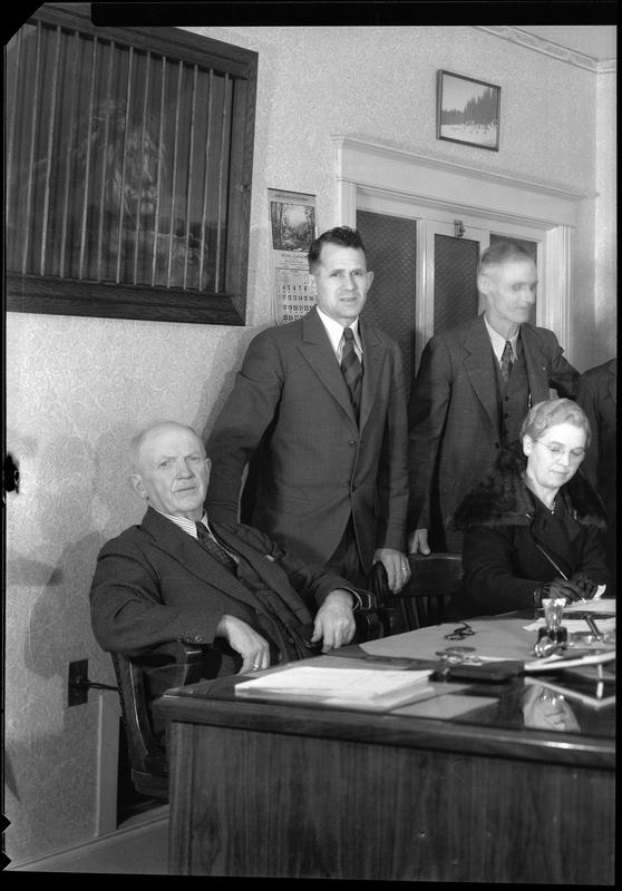 Two men are standing behind a seated man and woman. Two of the men are looking at the camera and the woman is writing something on the desk. A couple of paintings and a calendar hang on the wall behind them.