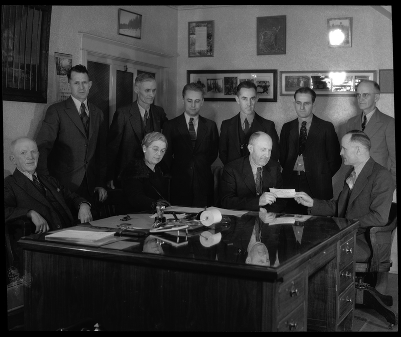 Photo of a group of men and a woman gathered around two men holding a check between them. Previously described as "Hatchery Photo (payment for land)," everyone is seated or standing around a large desk in a room. Various paintings are hung on the walls behind them.