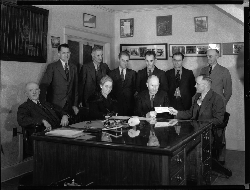 Photo of a group of men and a woman gathered around two men holding a check between them. Previously described as "Hatchery Photo (payment for land)," everyone is seated or standing around a large desk in a room. Various paintings are hung on the walls behind them.