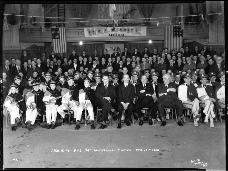Photo of a large group of men from the Fraternal Order of Eagles sitting with marching band members. Writing on the negative says: "Aerie No. 54 F.O.E. 34th Anniversary Meeting, Photo by Tabor's, Wallace, Idaho." There are American flags hanging in the background and a banner welcoming people to Aerie No.54.