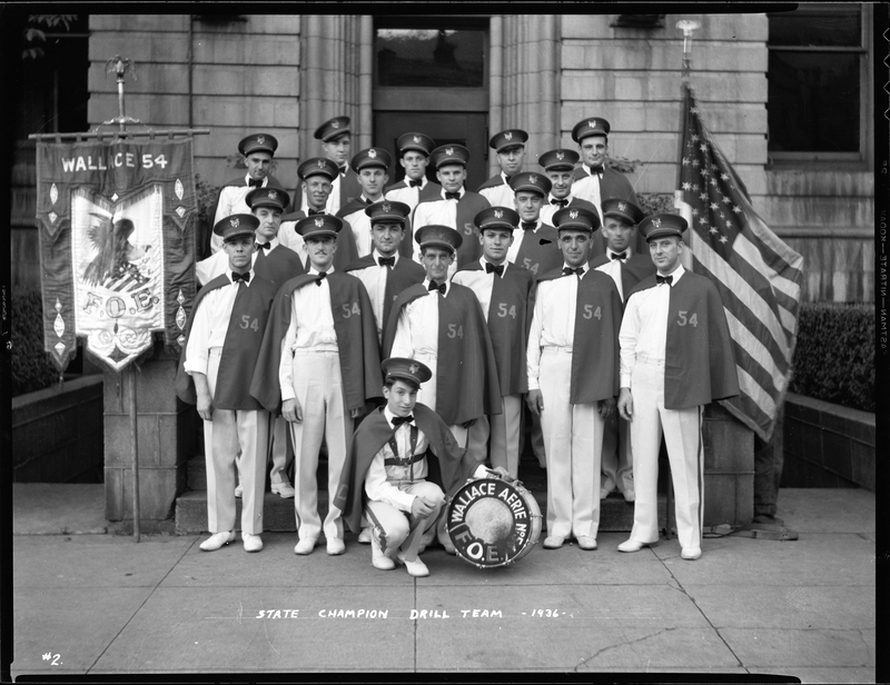 Group of men from the Fraternal Order of Eagles standing outside with a bass drum that has "Aerie No. 54 F.O.E." written on it. The negative has "State Champion Drill Team" written on it. The men are all wearing matching uniforms and there is a flag on either side of them.