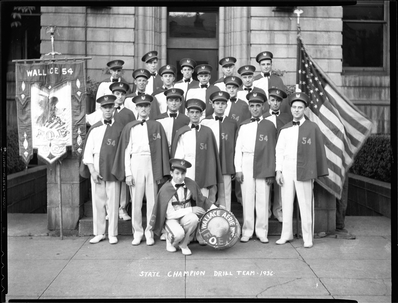 Group of men from the Fraternal Order of Eagles standing outside with a bass drum that has "Aerie No. 54 F.O.E." written on it. The negative has "State Champion Drill Team" written on it. The men are all wearing matching uniforms and there is a flag on either side of them.