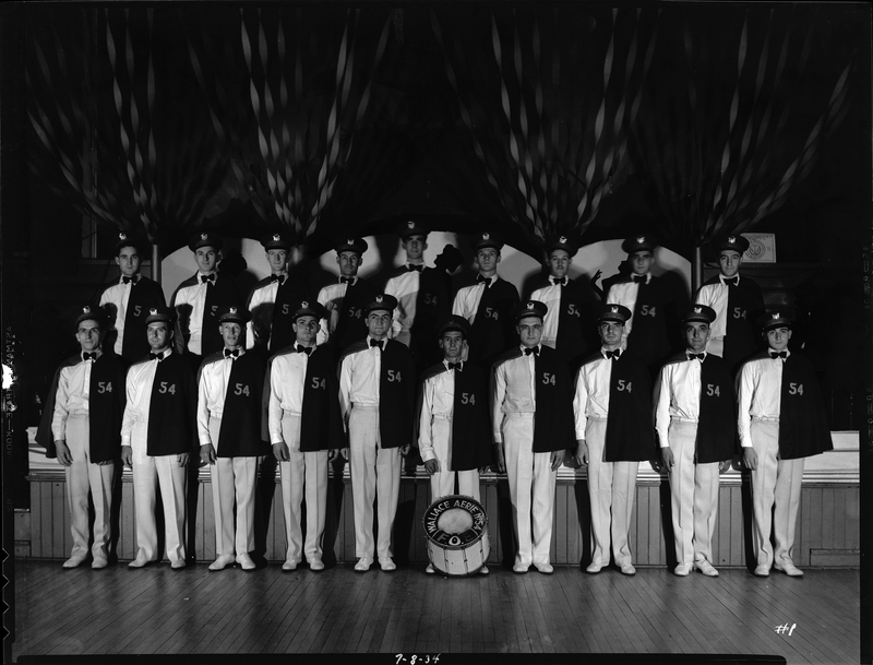 Group of men from the Fraternal Order of Eagles standing inside with a bass drum that has "Aerie No. 54 F.O.E." written on it. The men are all wearing matching uniforms with half of them standing on top of a stage behind the front row of men.