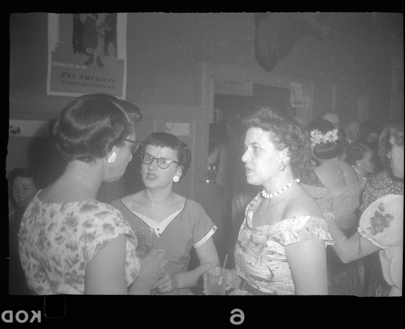 Three women at Rotary Party talking together. One woman has her back to the camera, and another is looking at the camera. They are holding drinks and wearing nice dresses.