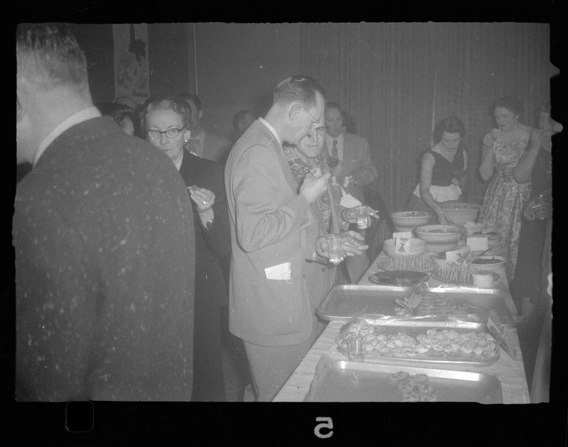 Photo of a table filled with food at Rotary Party with several people gathering food from it.