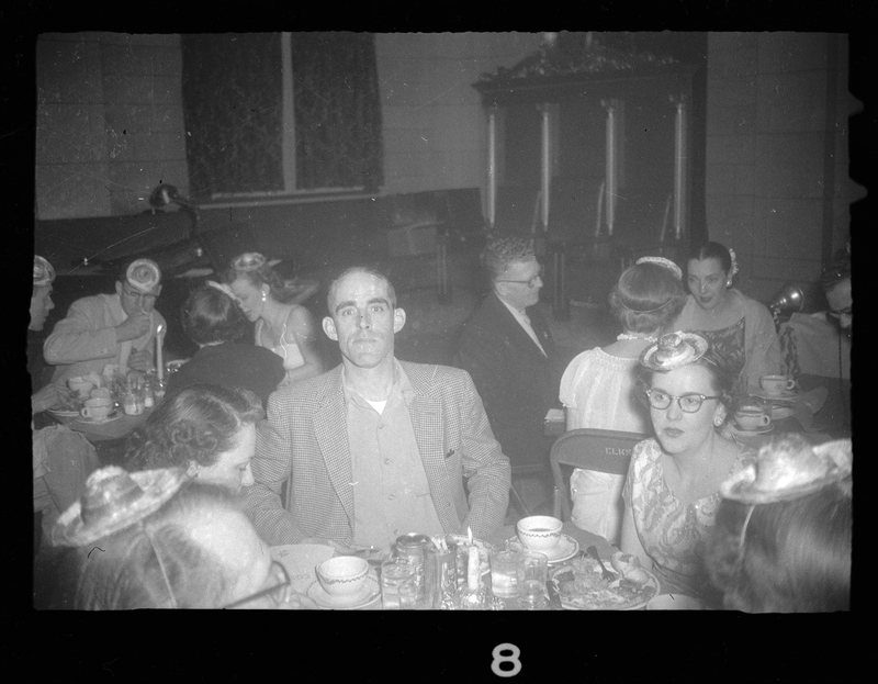 Photo of a man at Rotary Party. He sitting at a table with a group of women, and he is the only one looking at the camera. There are people at other tables in the background eating.