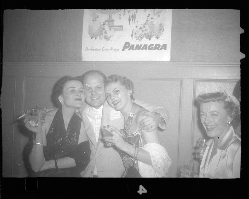 A man has his arm around two women with the third women standing off to the side. They are all dressed nicely, holding drinks, and smiling at the camera.