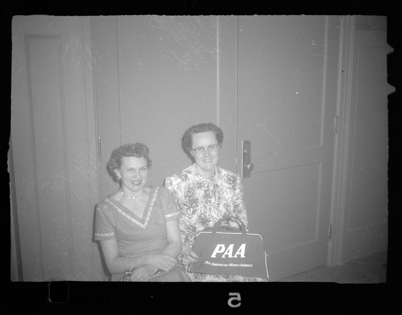 Two women sitting next to each other in front of a door at a Rotary Party. They are both smiling at the camera. One woman is holding a bag that reads "PAA. Pan American World Airways."