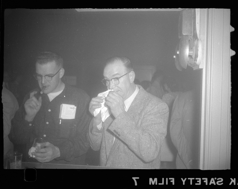 Two men stand next to each other at Rotary Party. One man is smoking a cigar and the other is wiping his nose.