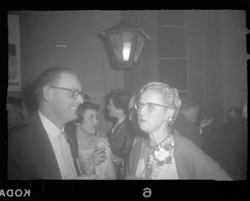 Photo of a man and a woman at a Rotary Party. The man is smiling at the woman. Other people can be seen in the background.