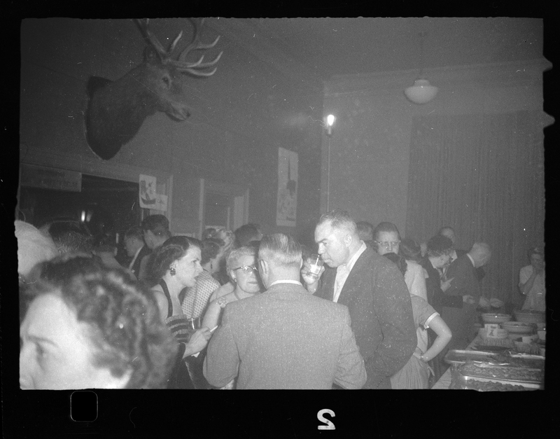 A small crowd of people mingling at Rotary Party. A buffet table of food can be seen in the corner, and there is the head of what is probably a deer mounted on the wall.