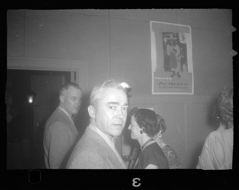 Photo of a man looking at the camera as the photo is taken. A few people can be seen behind him, as well as a poster on the wall.