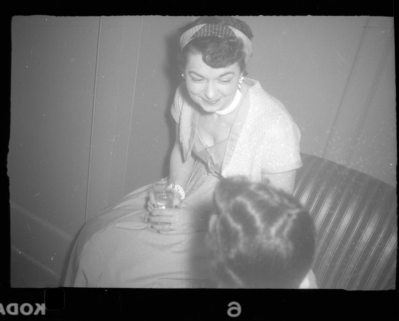 Woman sitting down at Rotary Party. The photo was taken at an angle, looking down at the woman. She is wearing a nice dress, holding a drink, and looking at someone who's back of head is visible.