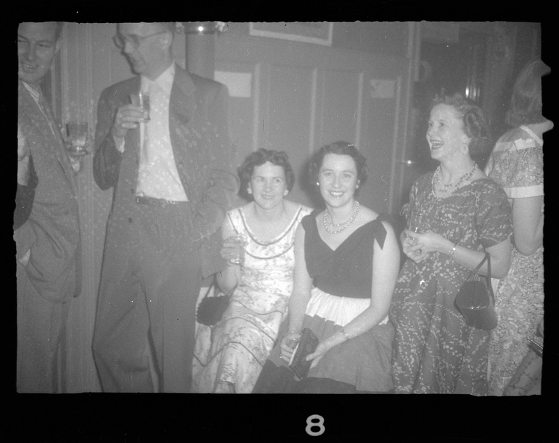 A small group of men and women at Rotary Party. Some of the women are looking at the camera, while the others are looking at each other. They are all well dressed and holding drinks.