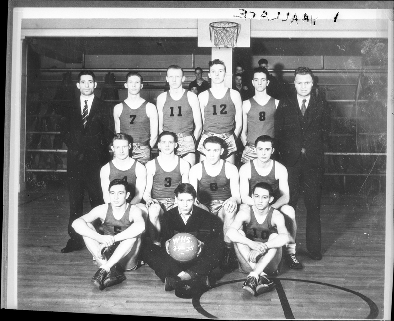 Photo of the Wallace High School basketball team members and presumably coaches gathered together in the gym. Some of them are wearing jerseys with numbers on them, and three others are wearing suits. One man is holding a basketball.