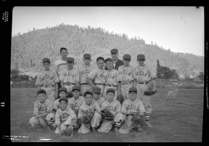 Group photo of the Hecla Little League baseball team and their coach. Front Row (4), from left to right: Roger Roth, Steve Bofenkamp, Sherm Ely, Mike Tattrie. 2nd Row (4): (unknown), Ralph Watson, (unknown), Bobby Walker. 3rd Row (7): Larry MacGuffie, Lee Lundal, Bob Sands, Larry Ohler, Larry Kiser, (unknown), (unknown). Back Row (2): (left) Coach George Vipperman and (right) Manager Art MacGuffie (father of Larry). They are wearing matching uniforms and a few are holding baseball gloves. (Possibly Hecla Mining).