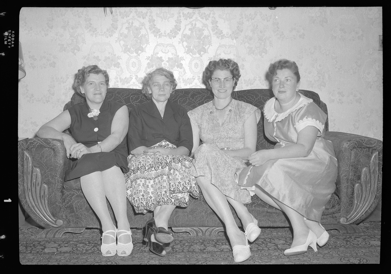 Four women sitting on a sofa together for photos. They are all wearing nice dresses.