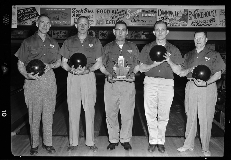 Photo of the five members of the Conoco bowling team. They are standing together in a bowling alley, four of them holding bowling balls and the man in the middle holding up a trophy.