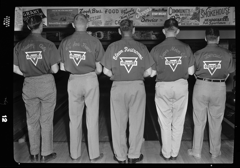 Photo of the five members of the Conoco bowling team. They are standing together, facing away from the camera to show the backs of their shirts, in a bowling alley.