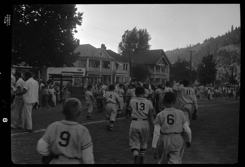 Photo of at least one little league baseball team walking away from the camera and towards a group of adults. Houses can be seen in the background.