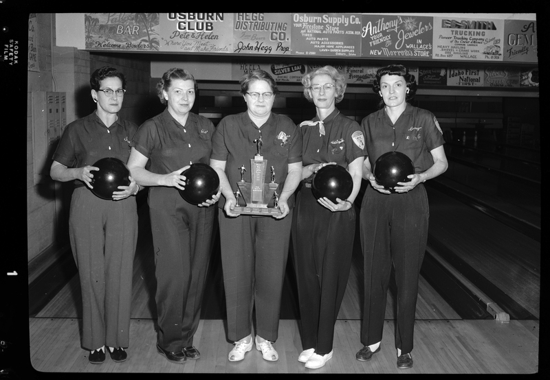 Photo of the Scoops bowling team posing together. Four of the five women are holding bowling balls and the fifth woman in the middle is holding a trophy.