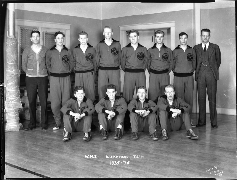 Photo of the Wallace High School basketball team and their coaches. The team is lined up in two rows. The front row of players are seated on the ground, while the back row are standing and everyone (aside from the coaches) are wearing matching uniforms.