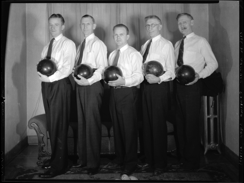 Five men from the City of Wallace bowling team are standing in a line. They are all wearing matching suits without jackets, and they are all holding bowling balls.