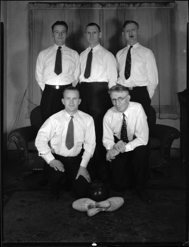 Five men from the City of Wallace bowling team are posing for a photo together. Two men are seated on a couch with the remaining three standing behind it. They are wearing matching suits without jackets and there are two bowling pins on the ground in front of the couch.