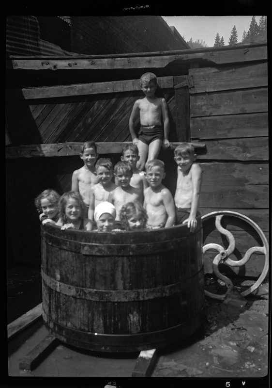 A group of kids are in a large barrel of water. Previously described as "kids in a tank at a Mine Yard in Burke, Idaho." Most of the children are standing up and they are all looking at the camera.