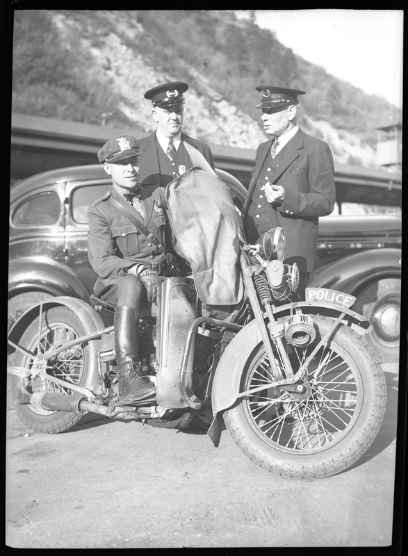 Three police officers are around a motorcycle, one sitting on it, and the other two standing around it. Previously described as "Tom Barrett, Vic Langley, and Charles Pugh. Police photographs."