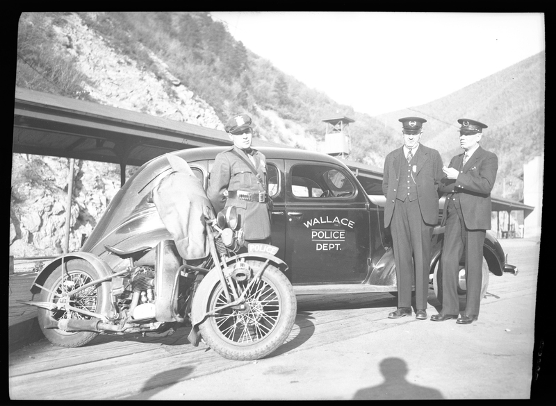 Three police officers are standing between a motorcycle and a car.They are in uniform and the car door has "Wallace Police Dept." on it. Previously described as "Tom Barrett, Vic Langley, and Charles Pugh. Police photographs."