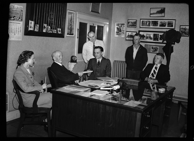 Five men and a woman are sitting a desk, looking at paperwork together. there are pictures and posters on the walls behind them. Previously described as "Ellis Hale in Coeur d'Alene Hardware Office."