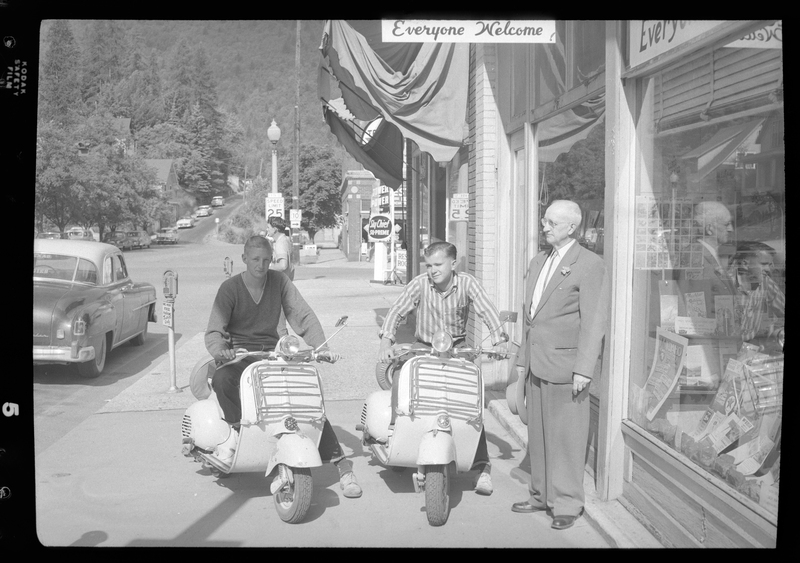 Two men sitting on motorcycles next to a man standing. They are parked on the sidewalk of a street and cars, trees, and shops can be seen in the background. Previously described as "Motorcyclists from San Francisco."