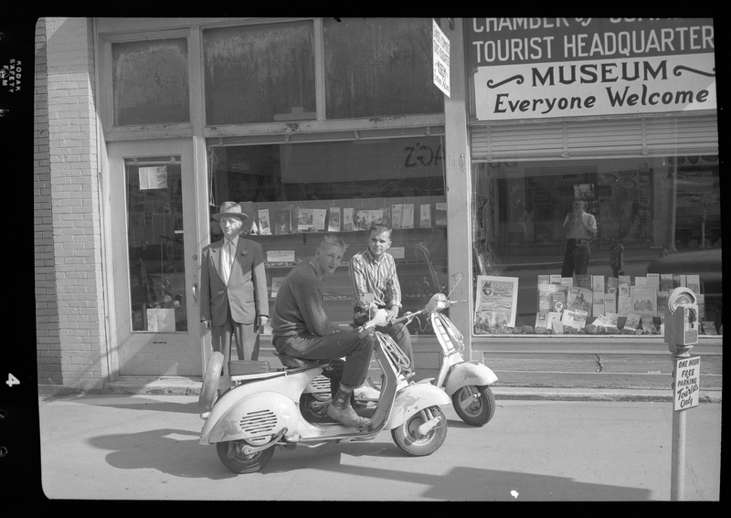 Two men sitting on motorcycles next to a man standing in front of a museum. They are parked on the sidewalk. Previously described as "Motorcyclists from San Francisco."