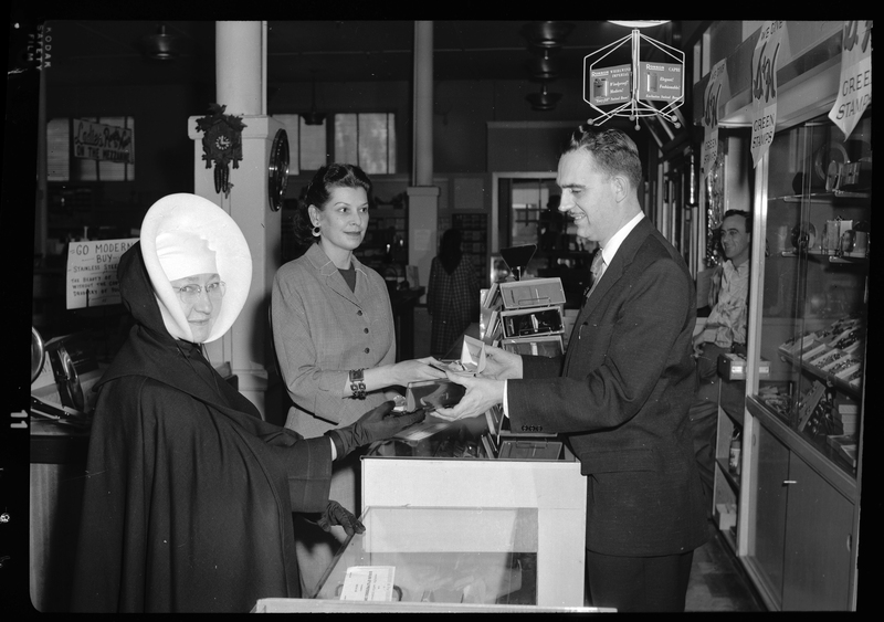 Two women, one who appears to be a nun, appear to be looking at jewelry at an unknown store. There is a man behind the counter showing them both a jewelry box. Previously described as "Anthony's Store."