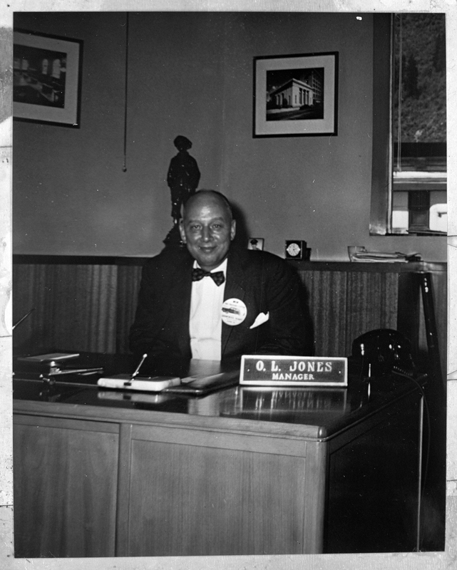 Photo of a man sitting behind a desk. There is a name placard on his desk that reads "O. L. Jones Manager."