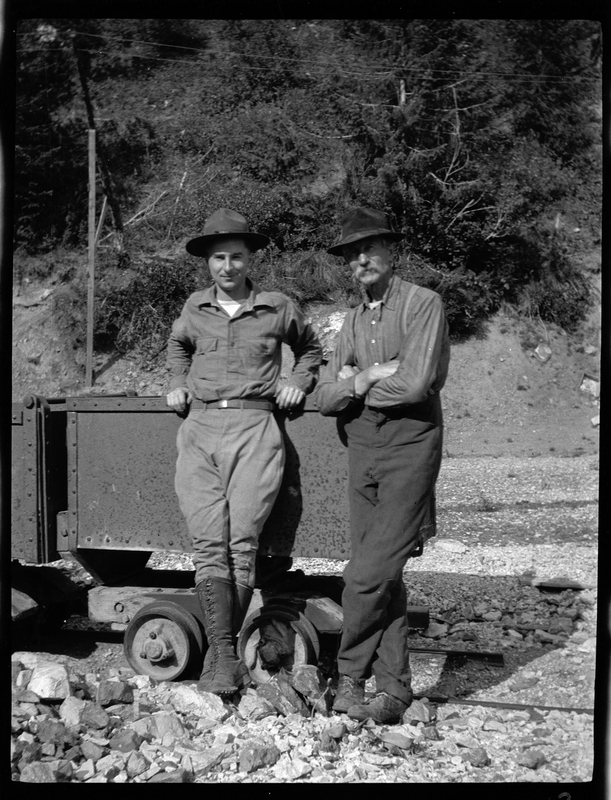 Photo of two unidentified men leaning against a cart outside. They are wearing similar clothes and looking at the camera.