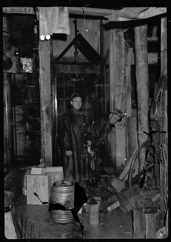 Photo of two unidentified men standing inside a building, probably some kind of workshop or storage space. They both have cigarettes in their mouths and are wearing hard hats with lights.