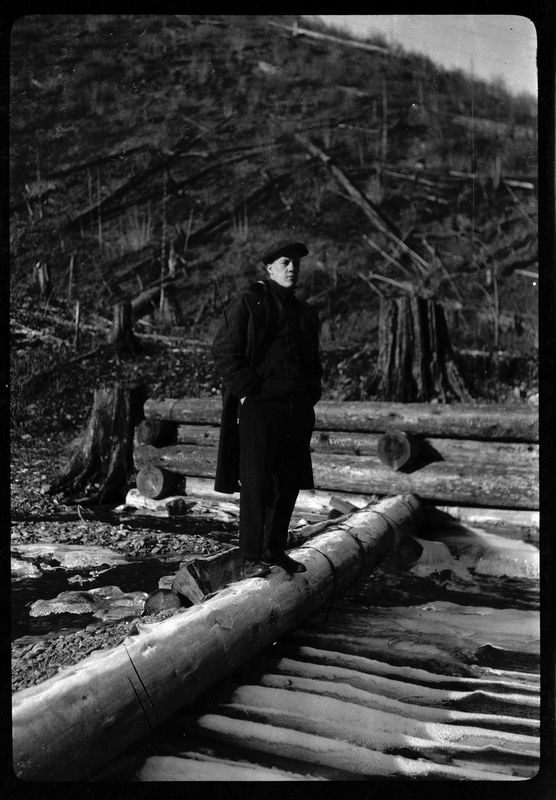 Photo of an unidentified man standing in what appears to be a logging yard. He is standing on a cut down log.