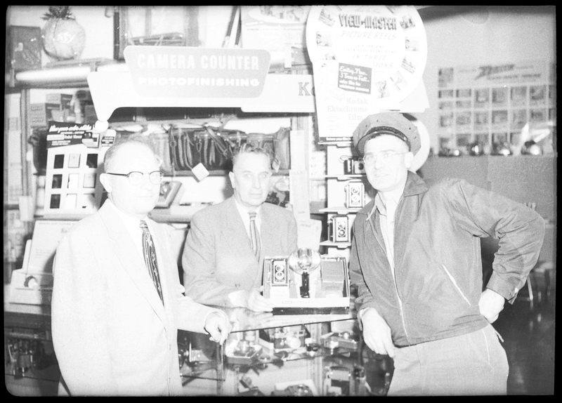 Three men standing inside of a store, one man possibly Lloyd Strope. 