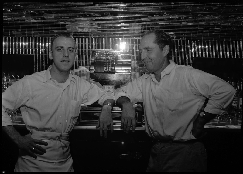 Harold Rullman and Babe Streeter, partners, standing in Sweet's Bar. They are standing next to each other behind the bar and leaning against the cash register.
