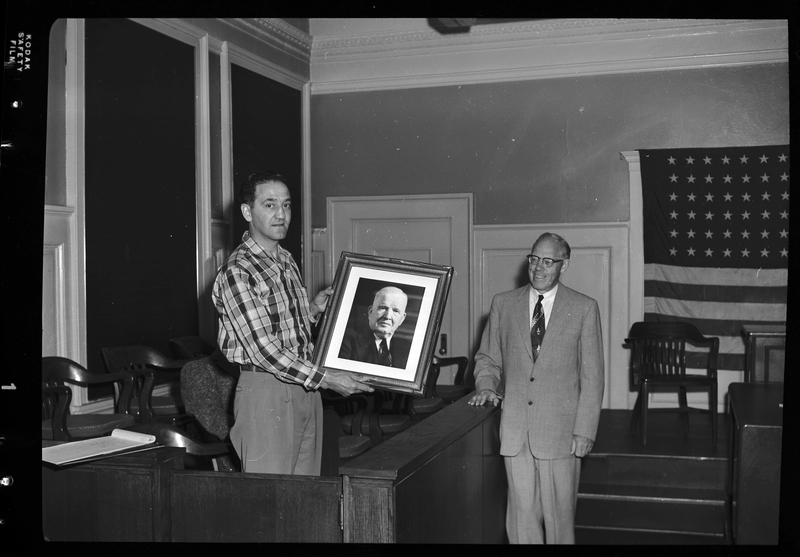 Two men standing together, possibly Shoshone County officials. One is holding a framed picture of Judge Featherstone. There is an American flag on the back wall.