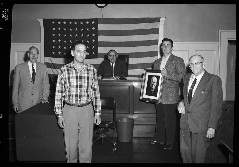 Five men together, possibly Shoshone County officials, and one is holding a framed photo of Judge Featherstone. One man is sitting behind a large desk and there is an American flag on the wall behind them.