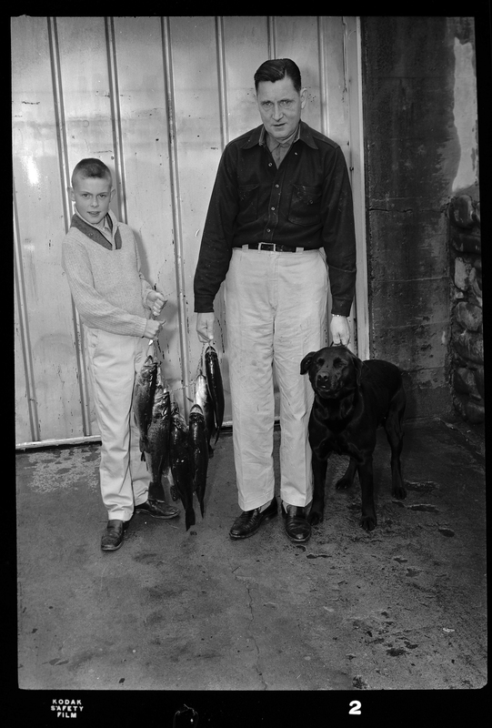 Les Randall with who is probably his son and his dog. They are holding a line of hanging fish between the two of them.