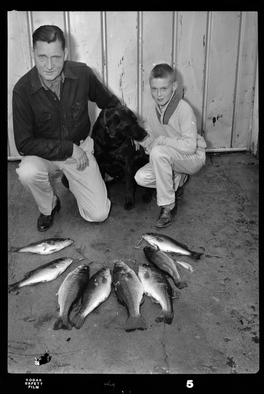 Les Randall crouched down with who is probably his son and his dog. There is eight fish on a line on the ground in front of them.