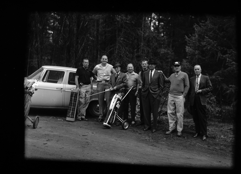Photo of eight men standing outside in front of a car. They have golf equipment, and they are all dressed well.